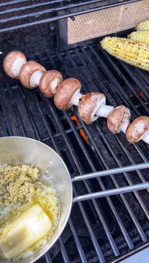 Brown mushrooms on a metal skewer on the grill with a small pot of melted butter and garlic in the forefront.