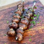 Two Grilled Garlic Butter Mushrooms on Skewers sitting on a wooden board. The mushrooms are garnished with flaky salt and fresh parsley.
