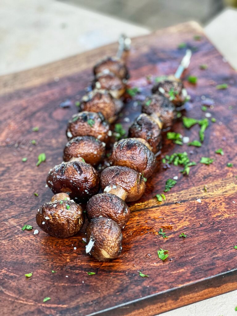 Two Grilled Garlic Butter Mushrooms on Skewers sitting on a wooden board. The mushrooms are garnished with flaky salt and fresh parsley.