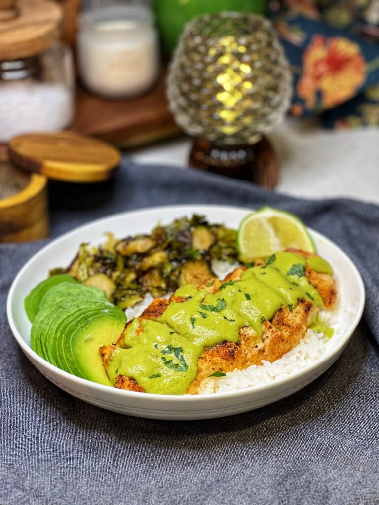 A white bowl holding roasted Brussels sprouts, sliced avocado and a bed of steamed rice that is topped with sliced chicken breast that is covered in a roasted poblano sauce. The sauce is bright green in color.
