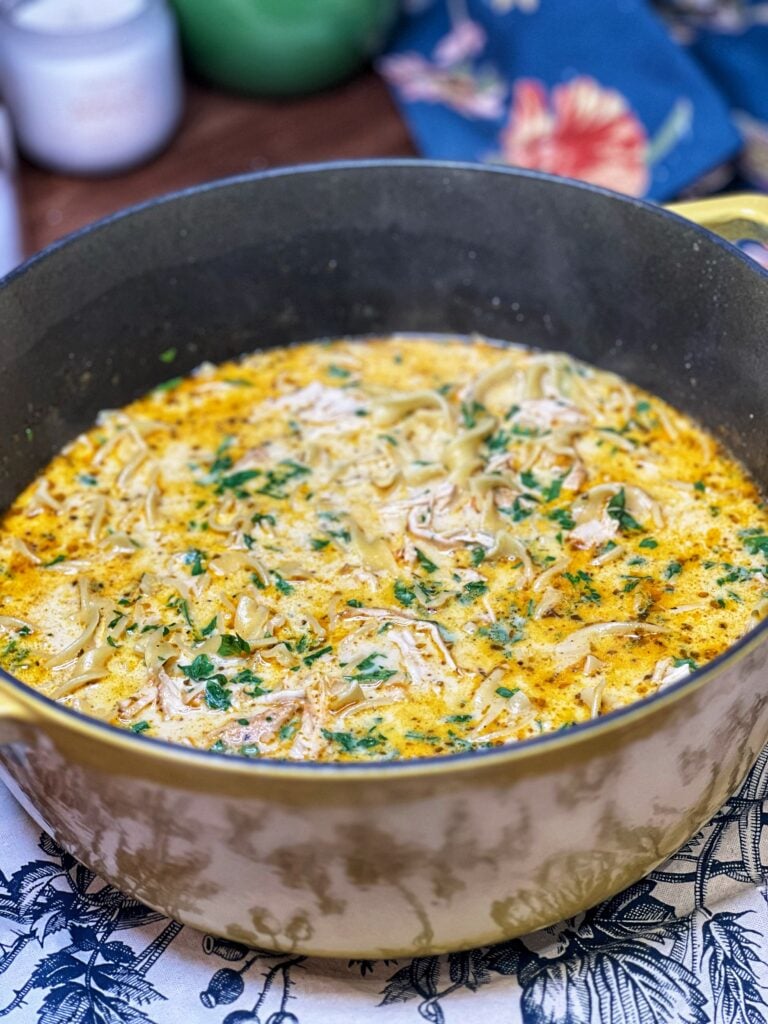A pot of creamy chicken noodle soup. The soup has a yellowish hue and is garnished with fresh parsley.