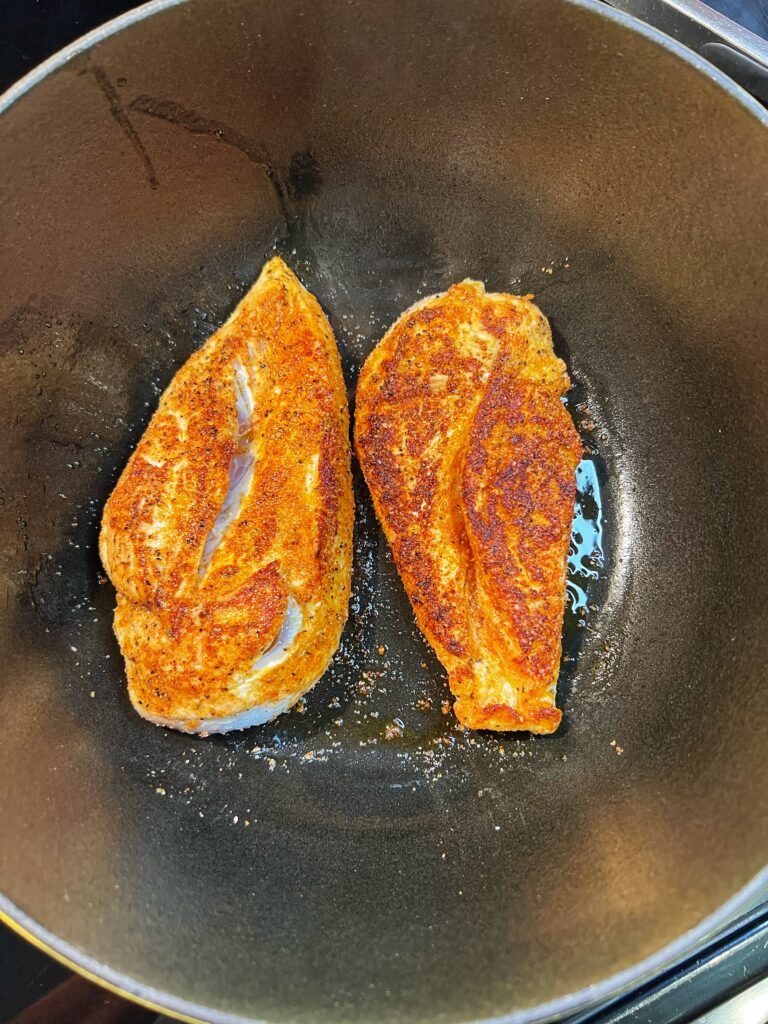 Two seasoned, boneless chicken breasts searing in a deep pot.
