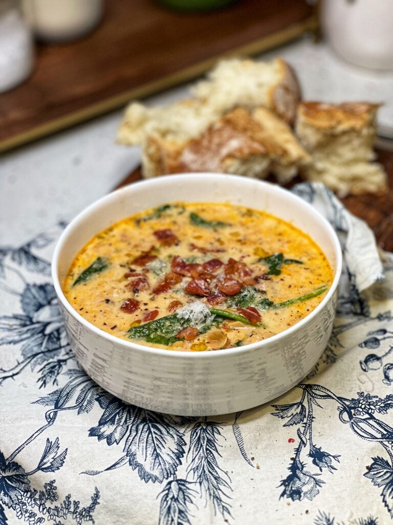 A white bowl filled with creamy tomato white bean soup. The soup has an orangish hue with spinach and is garnished with crisp diced bacon and grated Parmesan cheese.