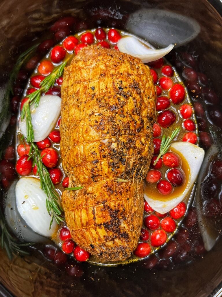 A seared boneless turkey roast in an oval, black crockpot surrounded by onions, whole cranberries, fresh rosemary sprigs in a liquid.