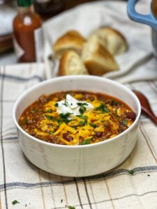 A white bowl filled with hearty chili with beans topped with shredded yellow cheese and sour cream.