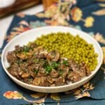 A white bowl filled with steak tips with mushroom gravy over rice with a side of peas.