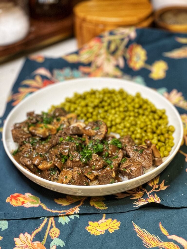 A white bowl filled with steak tips with mushroom gravy over rice with a side of peas.