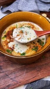 A brown bowl of she crab soup that is garnished with fresh parsley and paprika. The image is showing a wooden spoonful of the soup over the bowl.