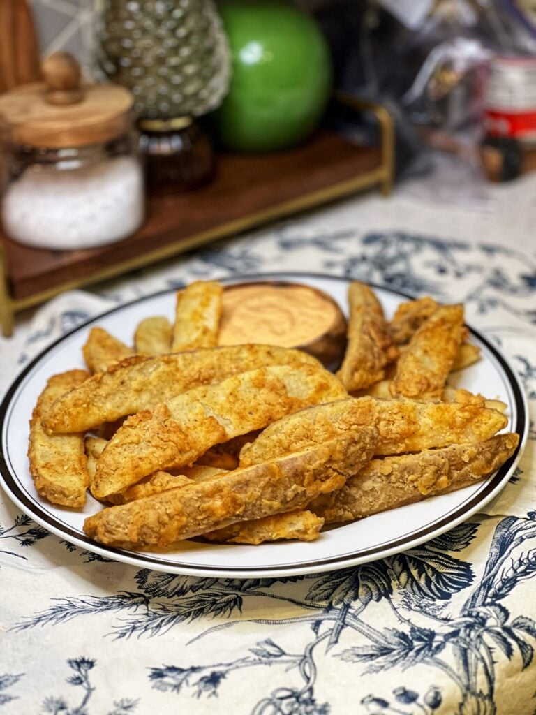A white platter of crispy potato wedges. The wedges are fried and served with a side of chipotle mayo.