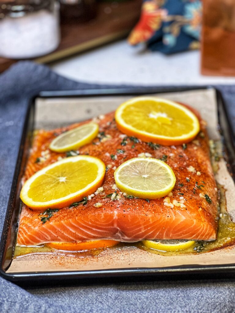 A large salmon filet sitting on top of sliced lemons and oranges on a baking sheet topped with an herb butter garlic sauce and more slices of lemon and orange.