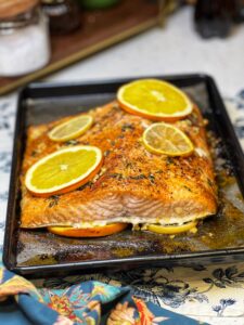 A small, black sheet pan holding Citrus Herb Butter Salmon filet. The salmon was baked on top of slices of orange and lemon and also topped with a few more slices.