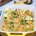 A yellow rectangular casserole dish filled with baked shrimp scampi. The pink shrimp are in a buttery sauce and topped with a browned breadcrumb topping. The dish is shown served with four lemon wedges and sprinkled with fresh chopped parsley.