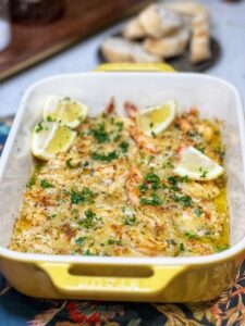 A yellow rectangular casserole dish filled with baked shrimp scampi. The pink shrimp are in a buttery sauce and topped with a browned breadcrumb topping. The dish is shown served with four lemon wedges and sprinkled with fresh chopped parsley.