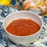 A white bowl filled with homemade cocktail sauce. The sauce is red and you can visually see flecks of ground horseradish. There is a small, white bowl in the background holding cooked shrimp.