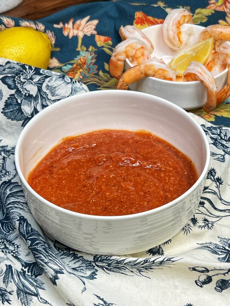A white bowl filled with homemade cocktail sauce. The sauce is red and you can visually see flecks of ground horseradish. There is a small, white bowl in the background holding cooked shrimp.