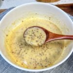 A white bowl of honey mustard dipping sauce. There is a wooden spoon in the bowl showing a spoonful of the sauce. The sauce has a yellowish hue and you can see the grainy bits of mustard.
