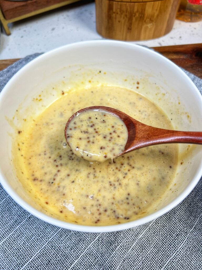 A white bowl of honey mustard dipping sauce. There is a wooden spoon in the bowl showing a spoonful of the sauce. The sauce has a yellowish hue and you can see the grainy bits of mustard.