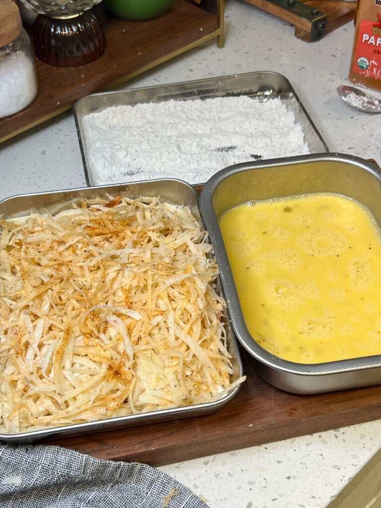 A dredging station for a potato crusted chicken recipe. One is filled with shredded, seasoned potatoes. The other is filled with beaten eggs, and the farthest one from the photo is seasoned flour.