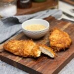 Two fried, potato crusted chicken breasts laying on a wooden cutting board. One is cut to show the inside. There is a small white bowl in the background filled with honey mustard dipping sauce.