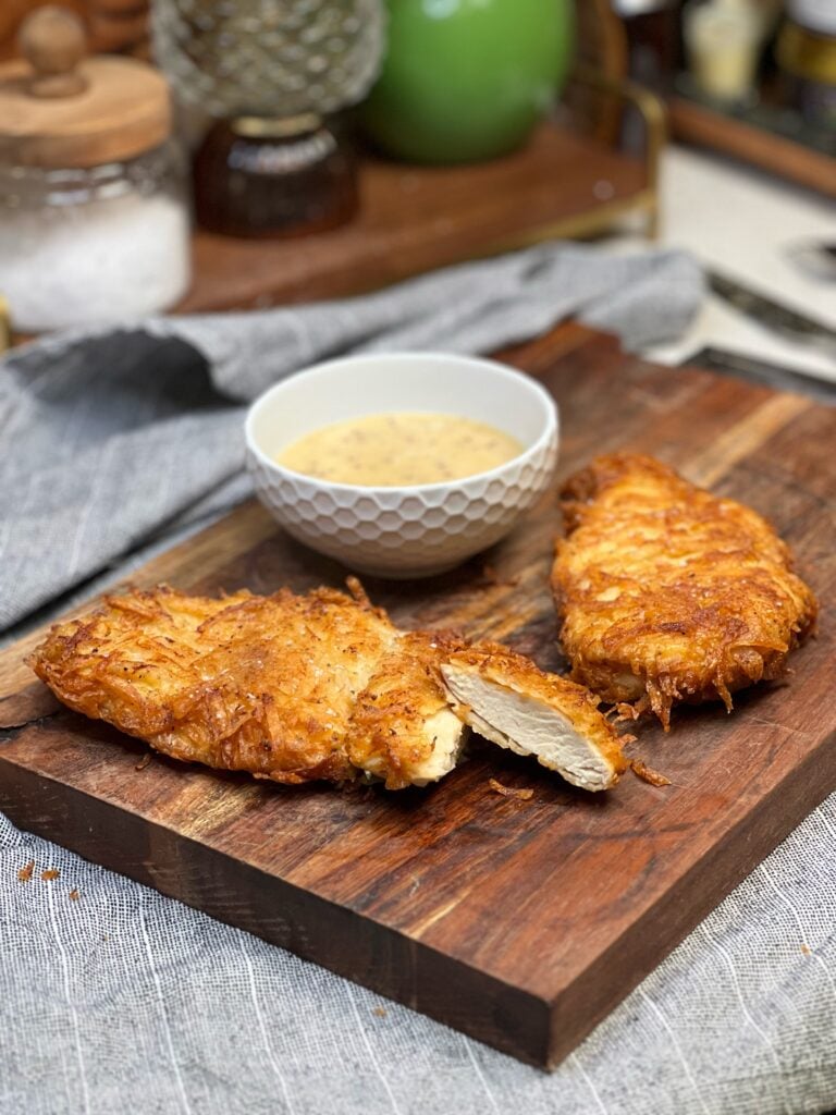 Two fried, potato crusted chicken breasts laying on a wooden cutting board. One is cut to show the inside. There is a small white bowl in the background filled with honey mustard dipping sauce.