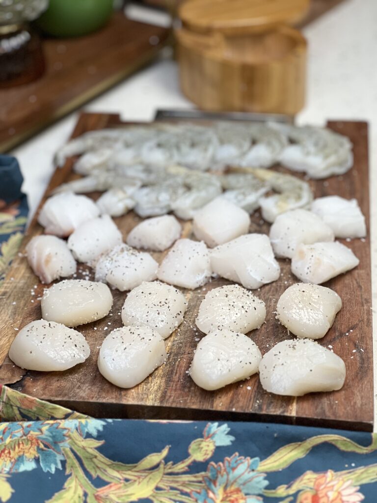 Fresh scallops, cod nuggets and shrimp laying on a wooden cutting board. The seafood is seasoned with salt and pepper.