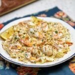 A white bowl holding creamy seafood pasta Alfredo. The dish features fettuccini noodles in a creamy, white seafood sauce with shrimp and scallops. It's garnished with a toasty breadcrumb topping, fresh parsley and served with lemon wedges.