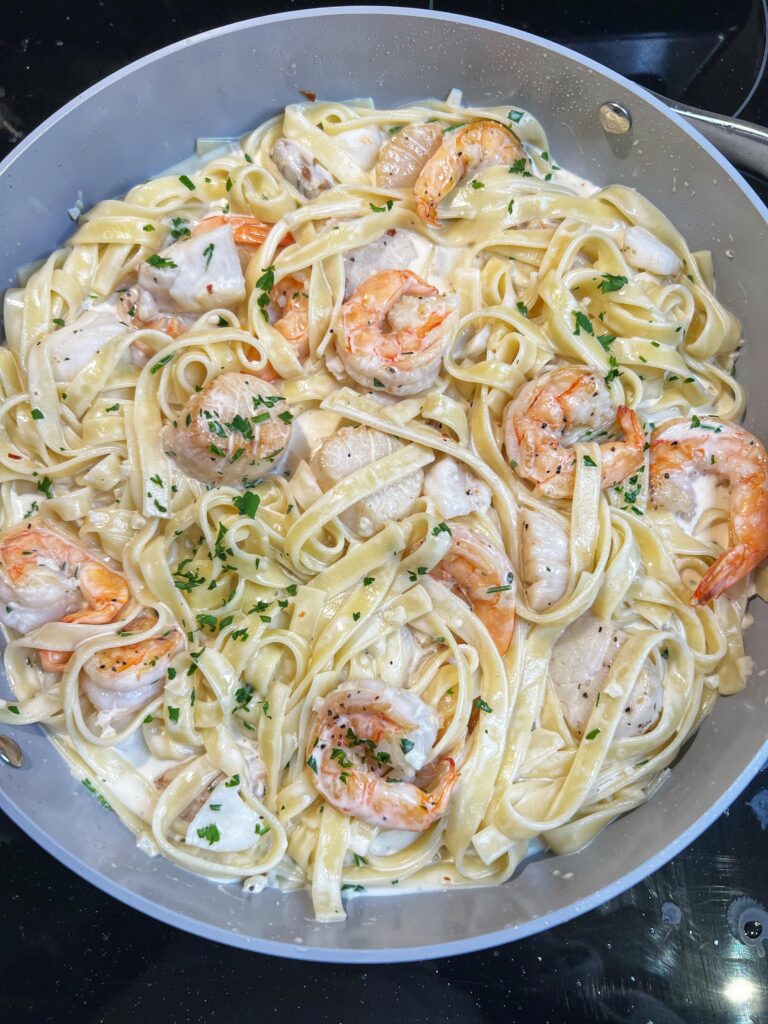 A pan of Creamy Seafood Pasta Alfredo. Fettuccini noodles are mixed with shrimp and scallops in a creamy sauce. The dish is garnished with fresh parsley.
