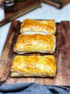 Three baked Ham and Cheese Turnovers sitting on a wooden cutting board. The turnovers are puffed up and golden brown.