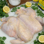 A raw, spatchcock chicken laying on a white plastic cutting board surrounded by halved lemons and fresh parsley. The chicken is laying flat.
