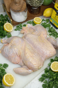 A raw, spatchcock chicken laying on a white plastic cutting board surrounded by halved lemons and fresh parsley. The chicken is laying flat.