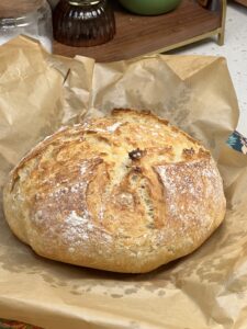 A loaf of simple no knead bread sitting on a piece of parchment paper. The round loaf of bread is golden and crispy.
