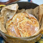 A loaf of Simple No Knead Bread sitting on top of a piece of brown parchment paper inside of a yellow Dutch oven.
