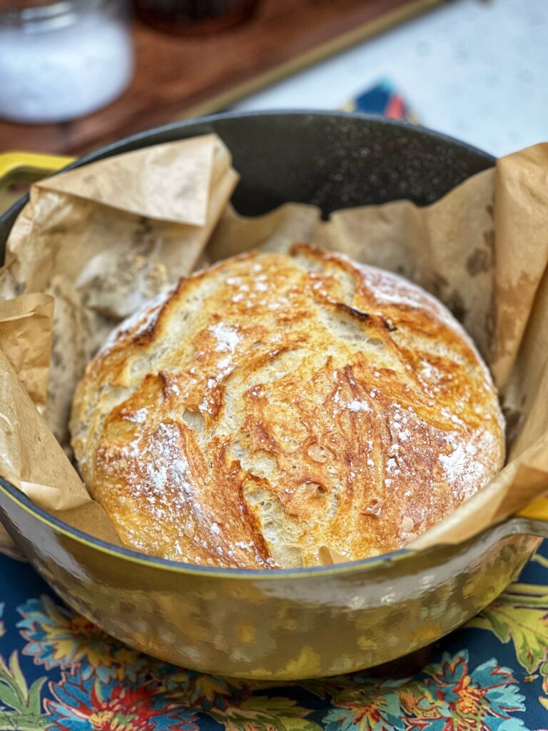 A loaf of Simple No Knead Bread sitting on top of a piece of brown parchment paper inside of a yellow Dutch oven.