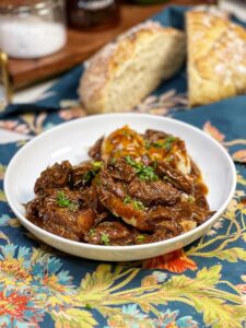 Two pieces of toast covered in shredded beef with gravy to create an open faced roast beef sandwich. The dish is served with mashed potatoes, which are also covered in gravy, and garnished with fresh chopped parsley.