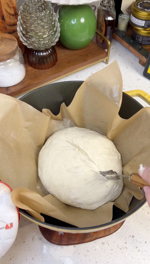 A ball of bread dough sitting on a piece of parchment paper inside of a yellow dutch oven. I am use a bread lame to score the dough.