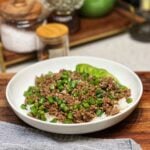 A white bowl of steamed rice covered in a ground beef and green bean stir fry. The green beans are diced and there is half of a sliced avocado in the back of the bowl.