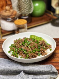 A white bowl of steamed rice covered in a ground beef and green bean stir fry. The green beans are diced and there is half of a sliced avocado in the back of the bowl.