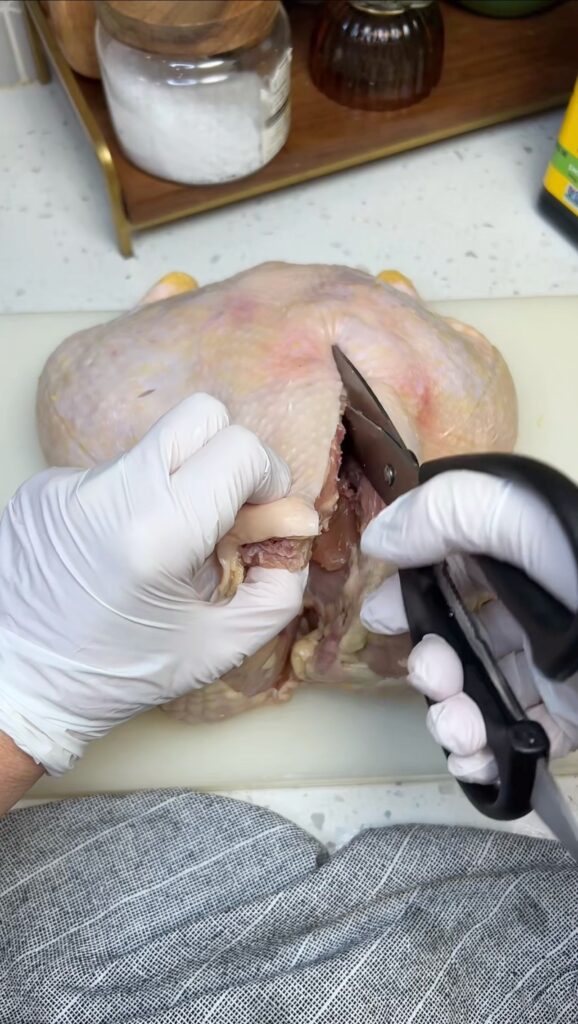 A whole chicken laying on a white plastic cutting board. I am using kitchen shears to cut out the backbone starting on the right side.