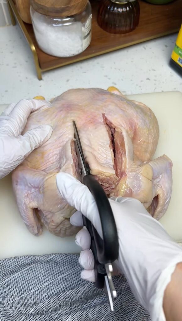 A whole chicken laying on a white plastic cutting board. I am getting ready to use kitchen shears to cut out the backbone on the left side. The right side is already partially cut.