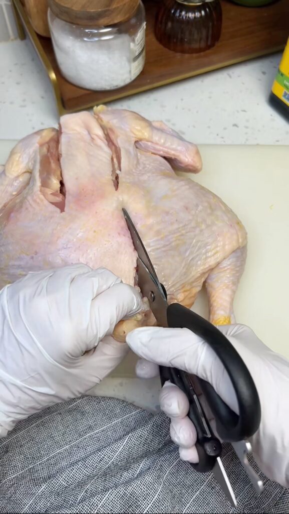 A whole chicken laying on a white plastic cutting board. I am using kitchen shears to cut out the backbone on the right side. I am almost through the side.