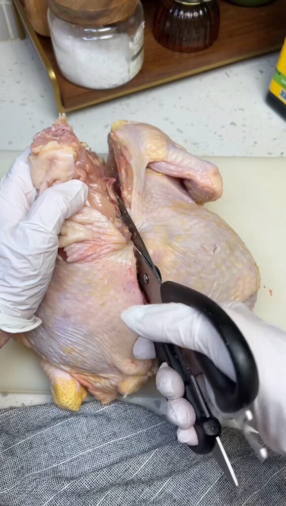 Cutting through the right side of the backbone of a whole chicken on a white cutting board.