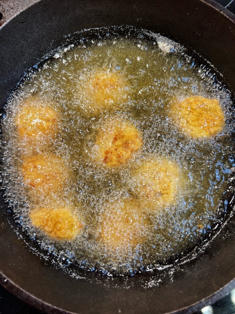 Eight breaded scallops frying in oil in a deep frying pot. 