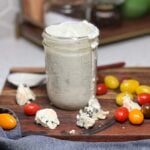 A mason jar filled with buttermilk blue cheese dressing. Some of the dressing is running down the side of the jar. The jar is placed on a wooden cutting board with cherry tomatoes and crumbled blue cheese.