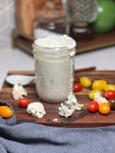 A mason jar filled with buttermilk blue cheese dressing. Some of the dressing is running down the side of the jar. The jar is placed on a wooden cutting board with cherry tomatoes and crumbled blue cheese.