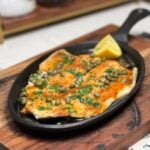 A filet of trout that has been baked then covered in a lemon butter garlic sauce with capers. The fish is sitting on top of a cast iron oblong pan.