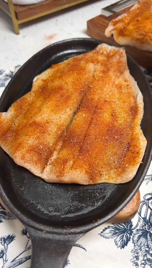 A butterflied, seasoned trout filet laying on an oblong cast iron pan.