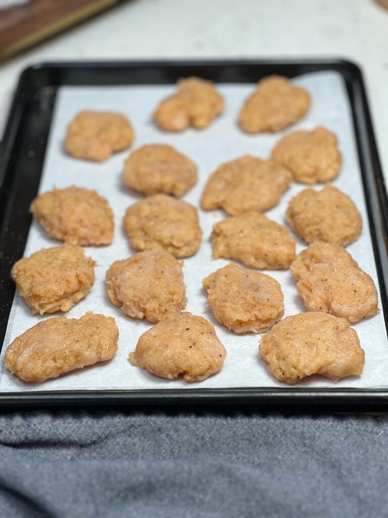Ground, seasoned chicken formed into nugget shapes laying on a sheet pan lined with parchment paper.