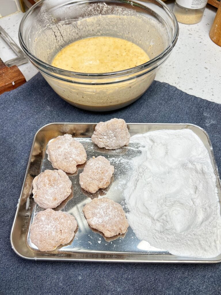 Chicken nuggets coated in rice flour laying in a stainless steel tray with a bowl of batter in the background.