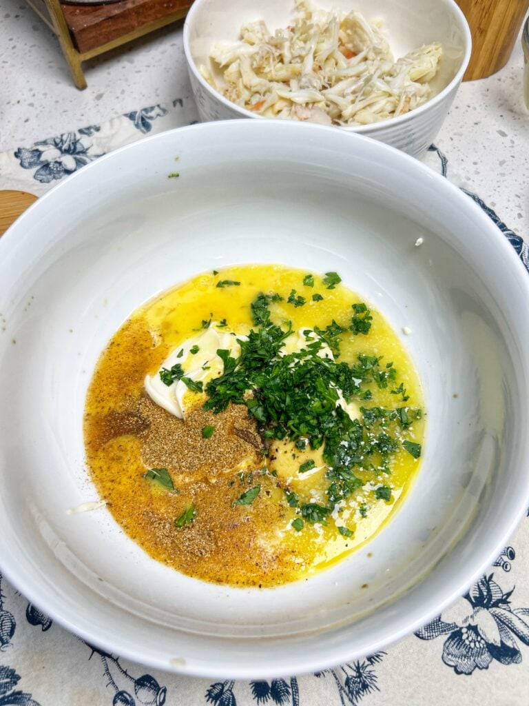 A white bowl of mayo, a beaten egg, seasonings and fresh parsley. It is unmixed.