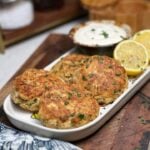 A white rectangular platter holding four canned tuna patties that have been sautéed and browned in olive oil. The patties are served with a bowl of lemon dill sauce and lemon wedges.